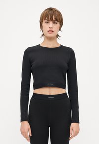 Svart croppet langermet topp laget av ribbestrikket stoff med rund hals og logobånd. Kombinert med matchende svarte høytaljede leggings.