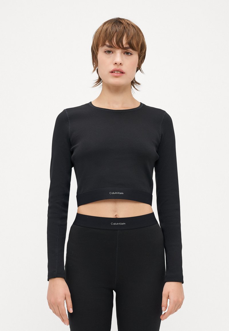 Svart croppet langermet topp laget av ribbestrikket stoff med rund hals og logobånd. Kombinert med matchende svarte høytaljede leggings.