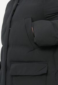 Chaqueta de plumas negra con una textura suave, mangas oversize y un gran bolsillo frontal. La tela parece lisa y duradera.
