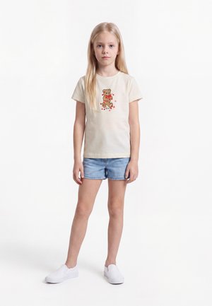 Jeune fille aux longs cheveux blonds portant un t-shirt beige avec un ours en peluche, un short en jean et des chaussures blanches slip-on, debout sur un fond blanc.