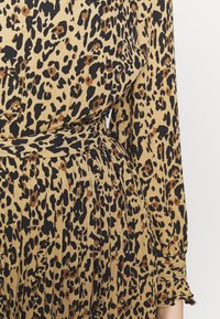 Robe à imprimé léopard en tan avec des taches noires, présentant une taille cintrée et une jupe plissée. Manches longues avec des poignets à volants.