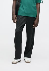 Μαύρο αθλητικό παντελόνι με φαρδιά εφαρμογή, κατασκευασμένο από ανοιχτό ύφασμα. Διαθέτει λευκές ρίγες στο πλάι και συνδυάζεται με λευκά αθλητικά παπούτσια.