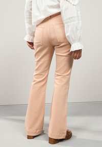 Rozkloszowane, jasnoróżowe spodnie z tylnymi kieszeniami i gładką fakturą, zestawione z białą bluzką z falbanianymi mankietami. Brązowe sandały na platformie.