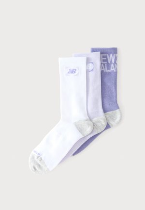 Trzy pary skarpetek New Balance do kostki w kolorach białym, jasnofioletowym i fioletowym z szarymi akcentami na pięcie i palcach, nałożone na siebie na jasnym tle.