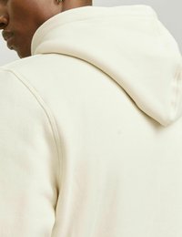 Světle béžová mikina s kapucí, vyrobená z texturované tkaniny, s viditelným šitím a volným střihem, s kapucí ležící vzadu.