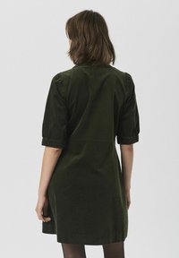 Mujer con pelo castaño hasta los hombros que lleva un vestido verde oscuro hasta la rodilla con mangas de largo hasta el codo, vista desde atrás.