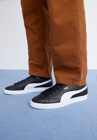 Sneakers in pelle nera con dettagli bianchi, suola piatta, punta rotonda e logo dorato. Abbinati a pantaloni marroni con risvolto su una superficie blu.