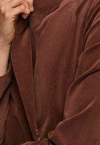 Hnědá kordurová bunda se strukturovaným povrchem a vertikálním žebrováním. Obsahuje překládaný límec a otevřený přední díl, což vytváří vrstvený vzhled.