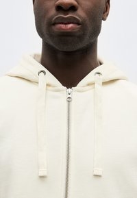 Nærbilde av en person som har på seg en kremfarget hettegenser med glidelås og snorer, som viser nederste del av ansiktet og nakken.