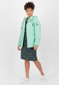 Veste imperméable vert menthe clair avec capuche, dotée d'accents noirs et de cordons de serrage ajustables, associée à une robe à motifs sombres et des baskets blanches.
