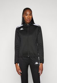 Svart zip-up jacka med huva, med vita detaljer på axlarna och logotyp på bröstet. Tillverkad av mjukt, elastiskt material.