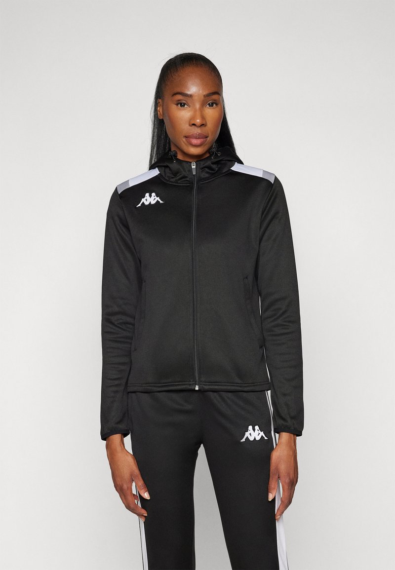 Svart zip-up jacka med huva, med vita detaljer på axlarna och logotyp på bröstet. Tillverkad av mjukt, elastiskt material.