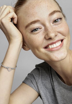 Silver charm bracelet with heart-shaped detail, featuring a textured finish. Model has light freckles and wears a gray short-sleeve shirt.