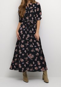 Robe maxi florale en tissu noir avec des motifs de fleurs colorées, fermeture à boutons sur le devant, manches courtes et détail de taille nouée. Présentée avec des bottes à talons beiges.
