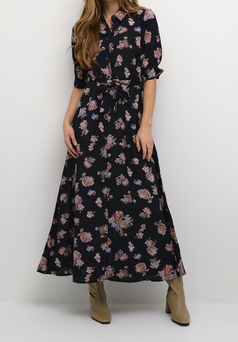 Robe maxi florale en tissu noir avec des motifs de fleurs colorées, fermeture à boutons sur le devant, manches courtes et détail de taille nouée. Présentée avec des bottes à talons beiges.