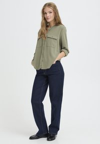 Chemise boutonnière olive avec deux poches poitrine, associée à un jean droit bleu foncé et des mocassins noirs. Tissu lisse, coupe décontractée.