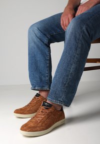 Braune Wildleder-Sneaker mit strukturierten Seitenflächen und cremefarbenen Sohlen, kombiniert mit blauen Denim-Jeans, aus der Kniewärts Sicht im Sitzen dargestellt.