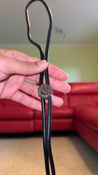 Μαύρο κορδόνι με πλεγμένη υφή, με εξάγωνη μεταλλική κλείδα σε γυαλιστερό ασημί φινίρισμα, κρατημένο ανάμεσα σε δάχτυλα μπροστά σε κόκκινο καναπέ.