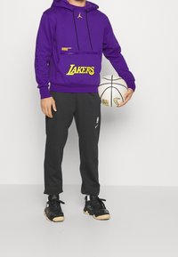Sudadera morada con el logo de "Lakers" en amarillo, bolsillo tipo canguro y capucha con cordón. Combinada con pantalones negros y zapatillas negras. Sostenido un balón de baloncesto.