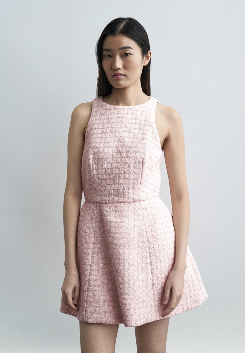 Νεαρή γυναίκα με μακριά σκούρα μαλλιά, φορώντας  αμάνικο, κοντό φόρεμα σε ανοιχτό ροζ με υφή, στέκεται μπροστά σε ένα απλό ανοιχτό γκρι φόντο.