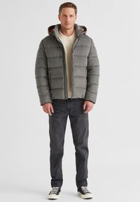 Graue steppjacke mit horizontalen Nähten, Frontreißverschluss, zwei seitlichen Taschen und Kapuze aus kontrastierendem Stoff. Getragen über einem leichten Pullover und Jeans.