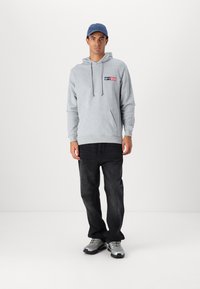 Grå hættetrøje med snore, frontlomme, logodetaljer. Båret med mørk denim jeans og grå sneakers. Blå kasket på hovedet.