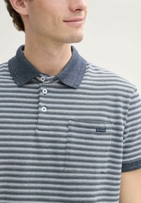 Gestreept poloshirt in marineblauw en wit. Voorzien van een gestructureerde marineblauwe kraag, drie knopen en een zak met een logo-tag op de borst.