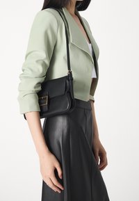 Bolso bandolera de cuero negro con un acento de herrajes dorados, llevado junto a una chaqueta verde menta y una falda de cuero negra. Textura suave.