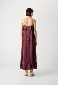 Bordeauxfarbenes Maxikleid mit rüschenschichten, dünnen Trägern und ausgestelltem Saum. Getragen mit beigen High-Heels. Glattes, strukturiertes Material.