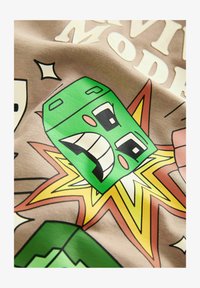 Grafik-T-Shirt mit einem grünen Cartoon-Charakter, der breit grinst, umgeben von explosiven Linien in Gelb und Orange auf einem beigen Hintergrund.