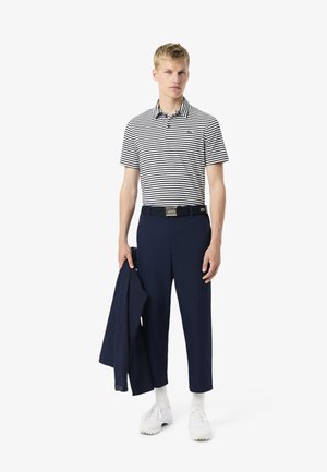 Gestreept poloshirt in marineblauw en wit, gecombineerd met marineblauwe broek en een zwarte riem. Model houdt een marineblauwe jas vast, draagt witte sneakers en sokken.