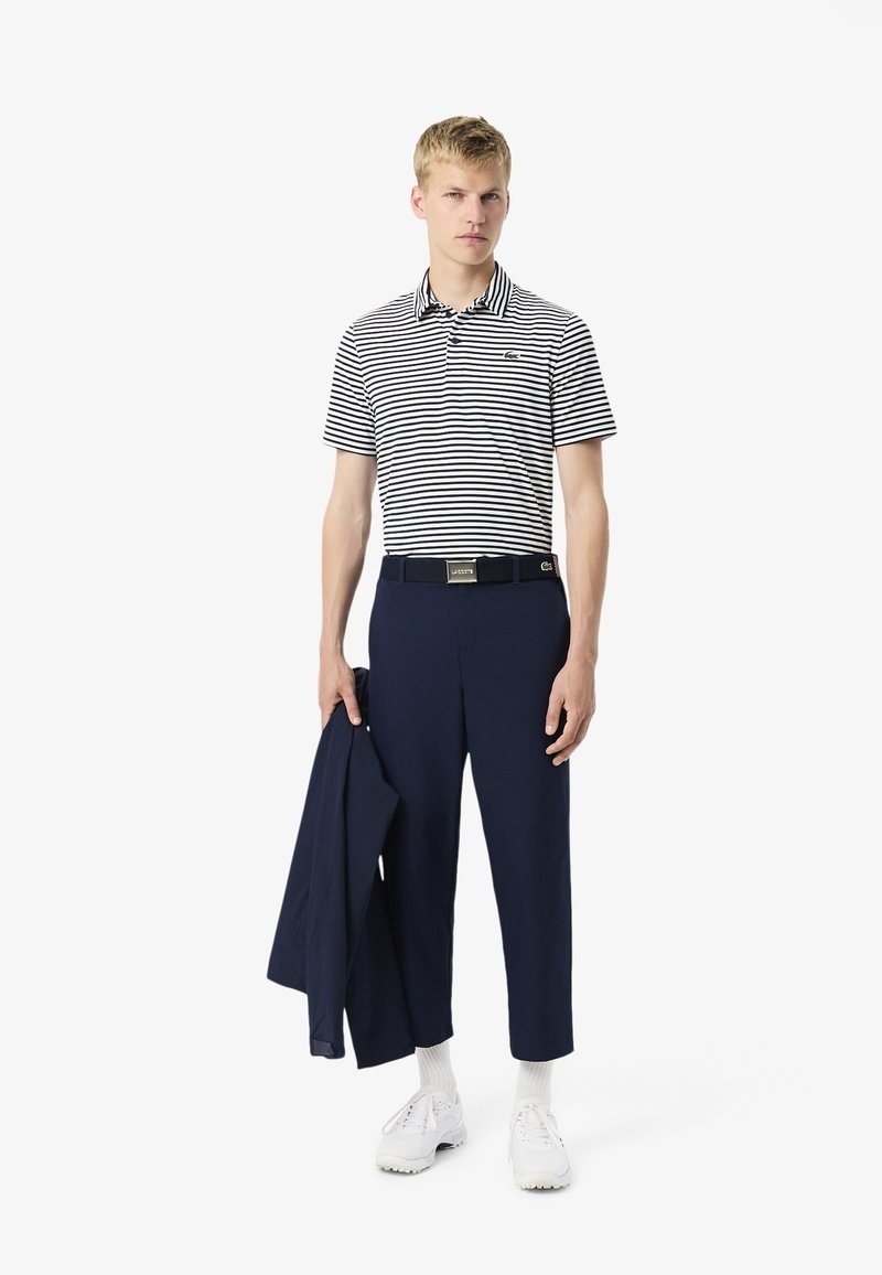 Polo à rayures en bleu marine et blanc, associé à un pantalon bleu marine et une ceinture noire. Le modèle tient une veste bleu marine, portant des baskets blanches et des chaussettes.