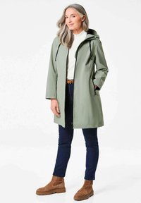 Lichtgroene waterdichte jas met capuchon, ritsen en zijzakken, gedragen over een witte top en donkere jeans. Bruine suède enkellaarzen.