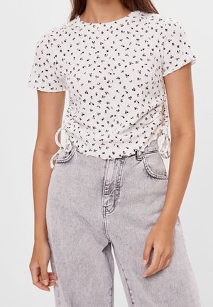 Hvid tekstureret top med sort blomsterprint, rund halsudskæring og sideskråning med bindebånd, kombineret med højttaljede lysegrå jeans.