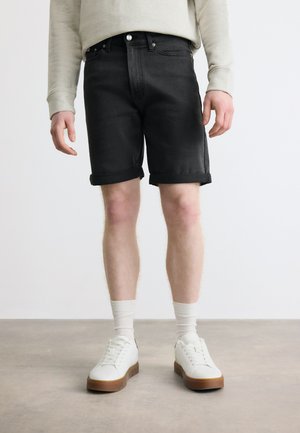 Muž v čiernych džínsových šortkách vyhrnutých, bielych ponožkách a bielych teniskách stojí na sivom podlaží pred bielym pozadím.