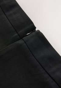 Zwarte stof met een gladde textuur, voorzien van een ritssluiting en nette naden. De focus ligt op het stiksel en de stofdetails.