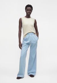 Mujer de pie con las manos en los bolsillos de pantalones anchos azul claro, vestida con top sin mangas beige y zapatos oscuros sobre un fondo liso.