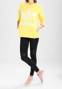 Gul hoodie med vit "Adidas"-logotyp, kombinerad med svarta leggings med meshpaneler och rosa textilsneakers.