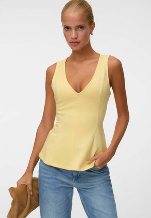 Mujer con cabello rubio, con una camiseta amarilla sin mangas con cuello en V y vaqueros azules, sosteniendo una chaqueta marrón en una mano.