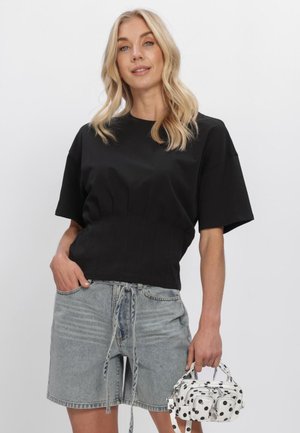 Blond vrouw met een zwart T-shirt met korte mouwen en lichte spijkershorts, die een kleine witte handtas met zwarte stippen vasthoudt.