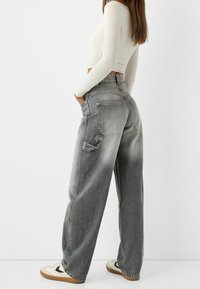 Nainen, jolla on yllään haalistuneet, leveät harmaat farkut suurella takataskulla, valkoinen pitkähihainen crop-top ja valkoiset kengät kumipohjilla.