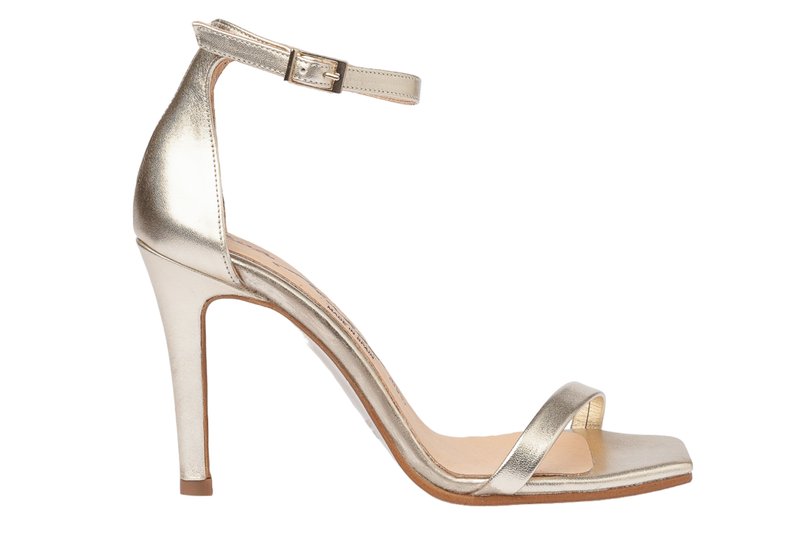 Sandal de tacón alto metálico dorado con correa en el tobillo y punta abierta. Textura suave, tacón delgado y diseño minimalista. Suela plana con ligero acolchado.