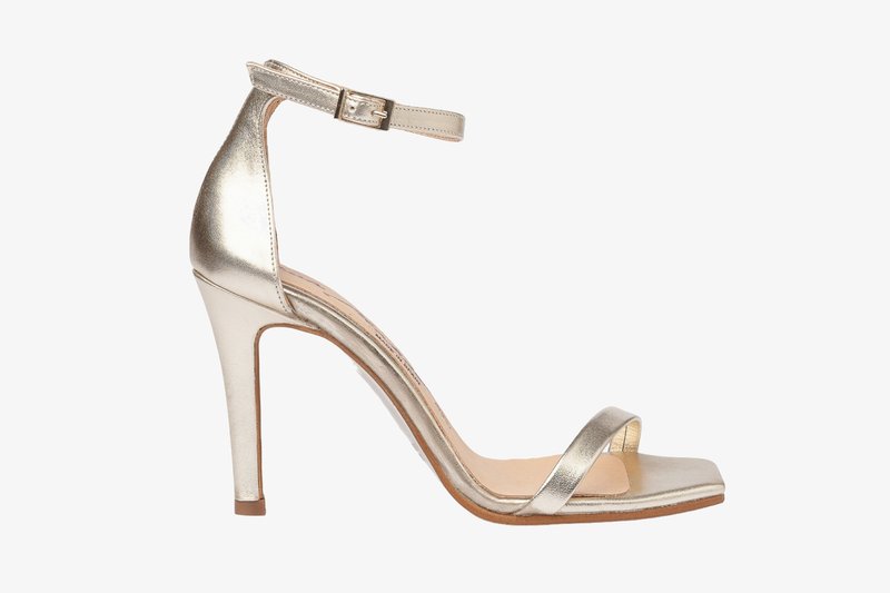 Sandal de tacón alto metálico dorado con correa en el tobillo y punta abierta. Textura suave, tacón delgado y diseño minimalista. Suela plana con ligero acolchado.