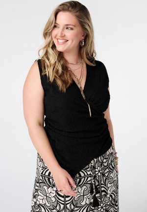 Lachende vrouw met golvend blond haar draagt een zwarte mouwloze top, gelaagde kettingen, hoepels en een zwart-witte rok met bloemenpatroon.