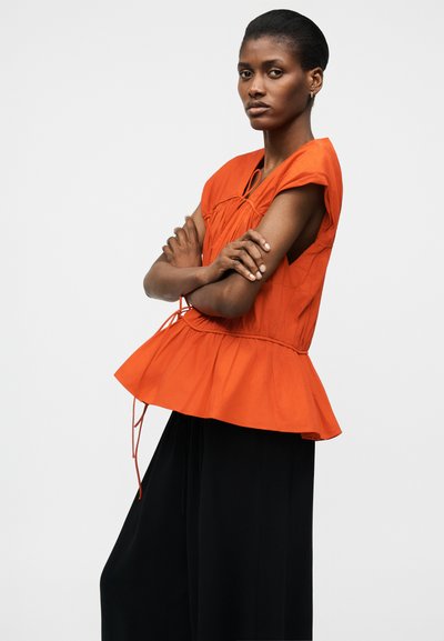 Naine, kellel on käed rüpes, seljas oranž varrukateta peplum-pluus ja laiad mustad püksid, seisab ühetaustalise hele tausta ees.