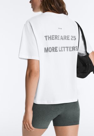 Kobieta w białej koszulce z napisem "THERE ARE 25 MORE LETTERS" na plecach i szarych szortach, trzymająca czarną torbę na ramieniu.