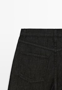 Schwarze Denim-Jeans Rückansicht, die den Bund, Gürtelschlaufen und eine einzelne Gesäßtasche mit sichtbaren Nähten auf weißem Hintergrund zeigt.