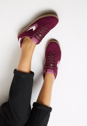 Bordeaux suede sneakers met witte accenten, platte veters en een lichtbruine rubberen zool. Opgerolde zwarte broekspijpen zijn gedeeltelijk zichtbaar.