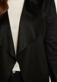 Veste noire en satin avec un devant ouvert et un col drapé, design superposé, portée sur un haut texturé beige clair. Bouton visible sur un pantalon foncé.