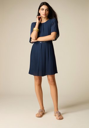 Donna con un vestito pieghettato blu navy a maniche corte, braccialetti d'oro e sandali nudi decorati, in piedi davanti a uno sfondo neutro.